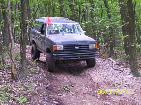 1995 Ford Bronco - A lifted dark green Ford Bronco with a roof rack, orange whip flag and oversized mud tires crawls down a rutted dirt trail through green su…