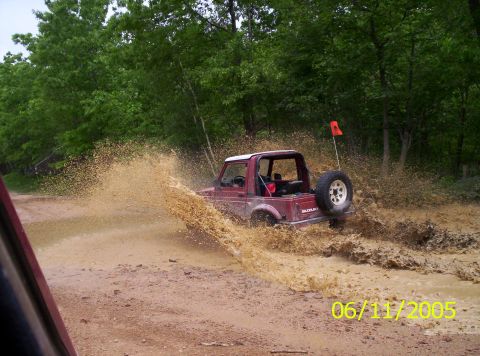 1995 Ford Bronco - A red Suzuki Samurai navigates a muddy trail, kicking up a large spray of water and mud.