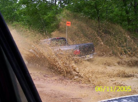 1995 Ford Bronco - A truck navigates a muddy obstacle, sending water and dirt flying.