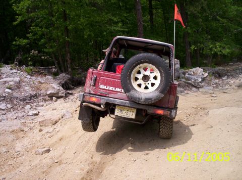 1995 Ford Bronco - Rear view of a topless maroon Suzuki Samurai 4x4 lifting its left rear wheel off a rocky dirt trail, with a swing-out spare on a white eigh…
