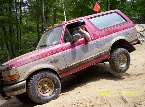 1995 Ford Bronco - Mud-splattered red-over-silver Ford Bronco descending a dirt bank, the driver leaning out the window flashing a peace sign. It wears Kumho…