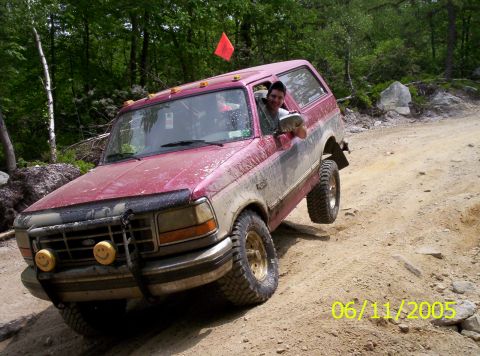 1995 Ford Bronco - Mud-splattered red full-size Ford Bronco leaning down a dirt trail bank with two occupants, the passenger hanging out the window holding a…