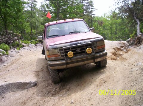 1995 Ford Bronco - A dust-covered maroon Ford SUV with a Ford-oval grille guard, twin yellow smiley-face driving light covers, roof cab lights and an orange t…
