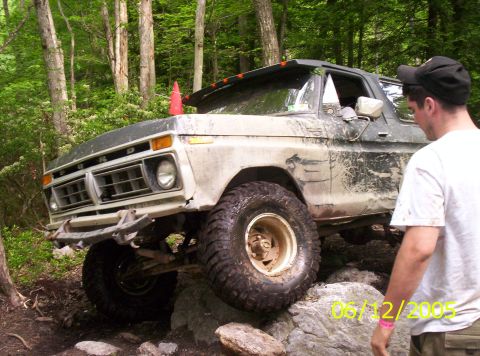 1995 Ford Bronco - A mud-caked 1970s Ford pickup with dent-side bodywork flexes over rocks on a wooded trail, front driver wheel drooped into a hole while the…