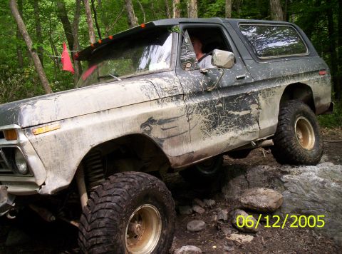1995 Ford Bronco - Mud-splattered white and dark-green late-1970s full-size Ford Bronco flexing over rocks on a wooded trail, driver visible through the open…