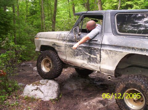 1995 Ford Bronco - A mud-splattered dark blue full-size Ford Bronco crawls over rocks on a wooded trail, front end angled up, with a man leaning out of the dr…