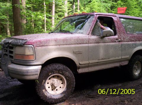 1995 Ford Bronco - Two-tone maroon-over-silver Ford Bronco caked in dried mud, shown from the driver's side on a muddy wooded trail with the driver's arm rest…
