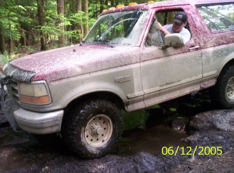 1995 Ford Bronco - A red-over-white Ford Bronco caked in mud dropping into a wooded mud hole, shot from the front left with the driver leaning out the open wi…