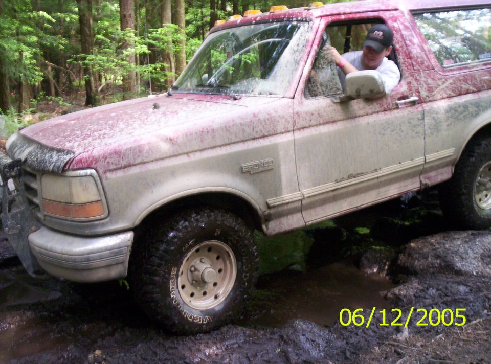 Muddy red and white Ford Bronco in a mud hole with driver leaning out the window