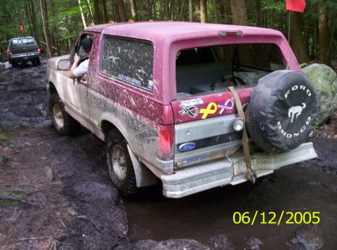 1995 Ford Bronco - Three-quarter rear view of a mud-splattered red-over-white Ford Bronco working through a wet rocky trail in the woods, date-stamped 06/12/2…