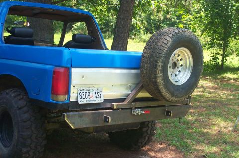 1987 Ford Bronco - Rear view of a blue 1987 Ford Bronco, showing its spare tire and custom bumper.