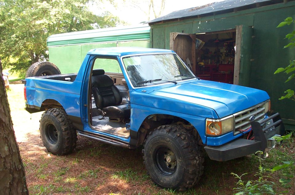 Blue Ford Bronco with aftermarket bumper and large tires, driver's door open, spare tire mounted in bed