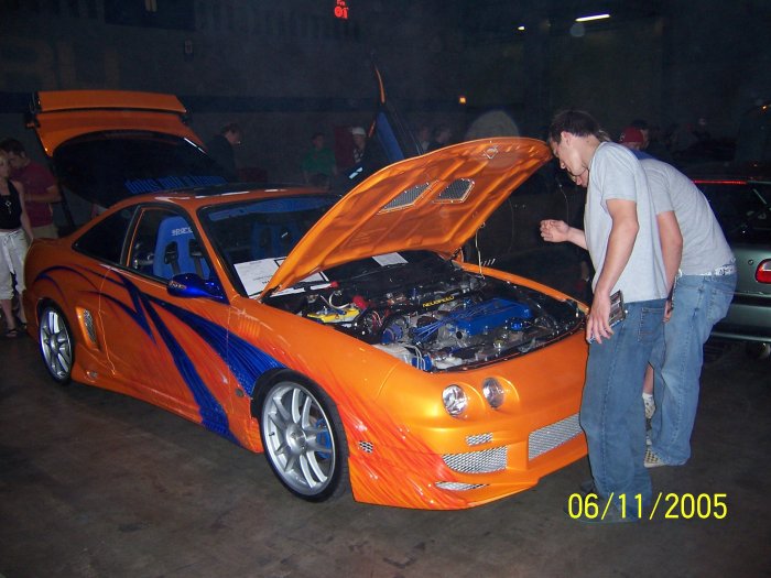 Orange sports car with blue flame stripes, hood open revealing engine, two people looking at it