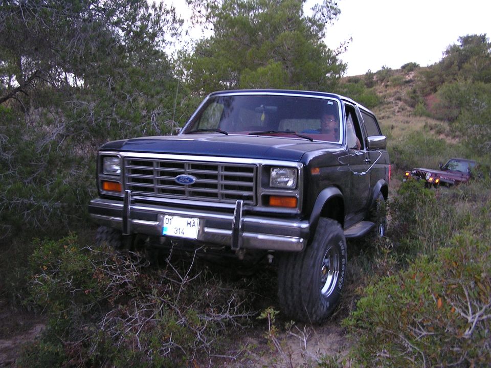 Dark blue lifted Ford Bronco with brush guard and off-road tires in scrub brush; Jeep in background