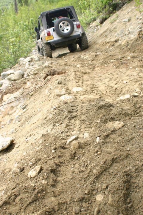 2003 Jeep TJ - A silver Jeep TJ navigates a steep, rocky dirt trail, kicking up dust as it climbs.