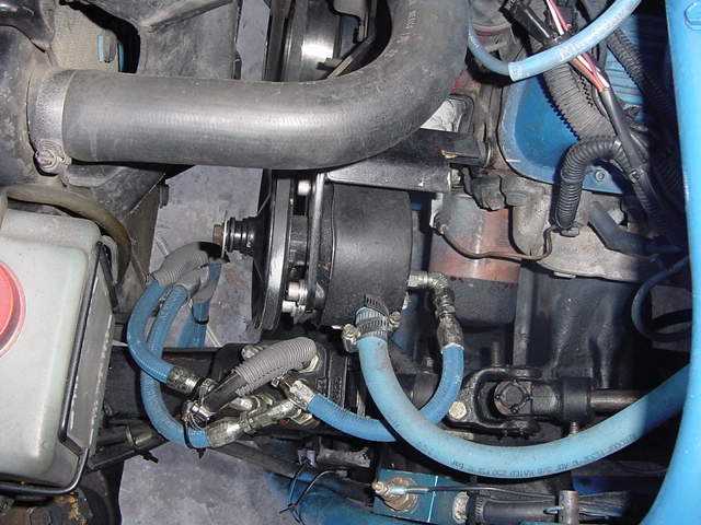 Engine bay with hoses, belts, and engine block visible. Blue tubing and black components dominate the frame