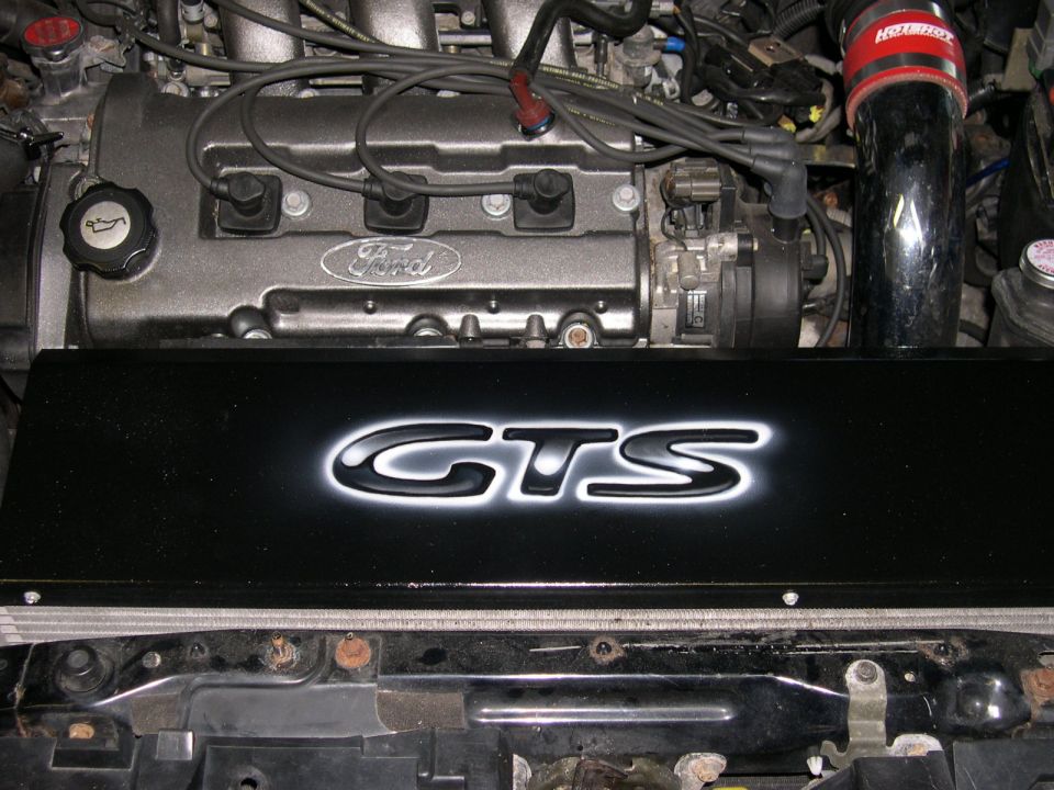 Close-up of a Ford engine with a black cover that says GTS. There's a red Hotchkis air intake tube