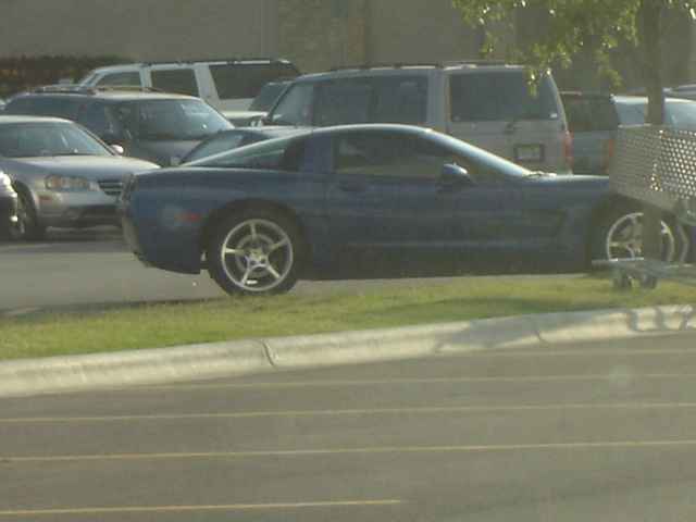 Dark blue Corvette coupe in side view parked on grass with a busy parking lot behind it