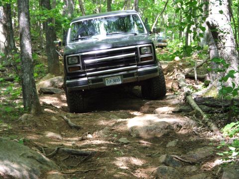 1996 Ford Bronco - A dark SUV navigates a challenging rocky trail surrounded by dense forest.