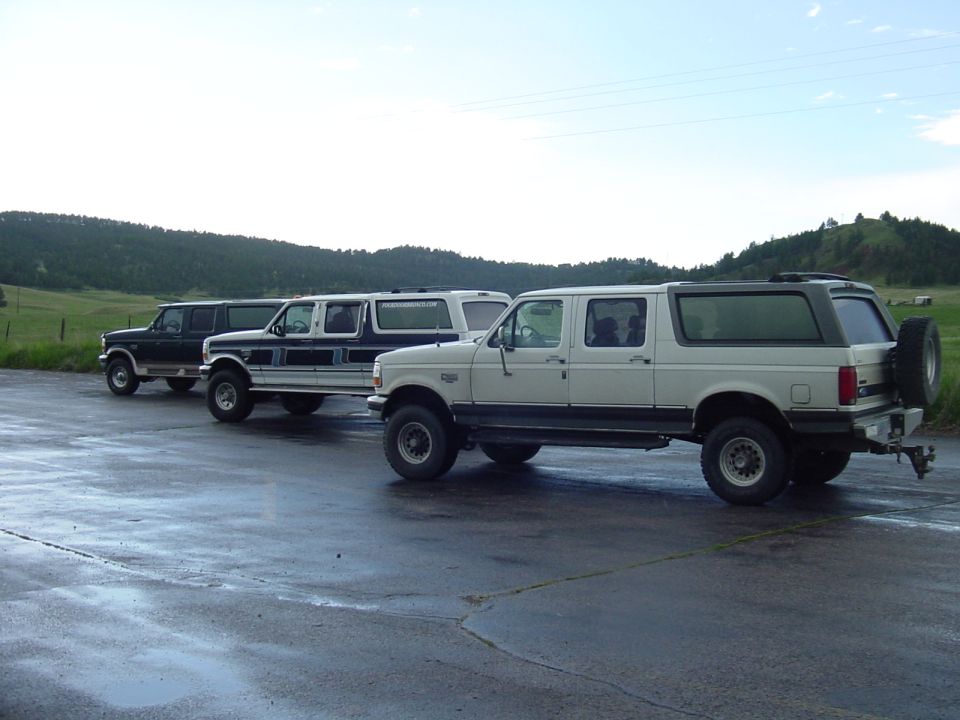 Three lifted full-size Ford trucks and SUVs lined up in a wet parking lot with hills behind