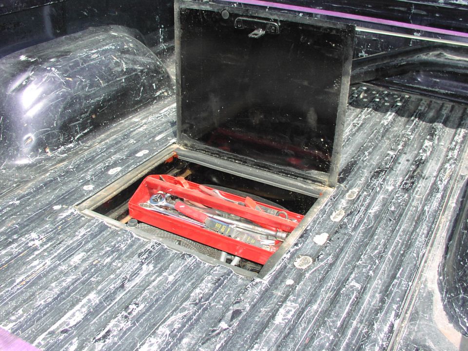 Truck bed with open black storage compartment revealing a red tool case with wrenches