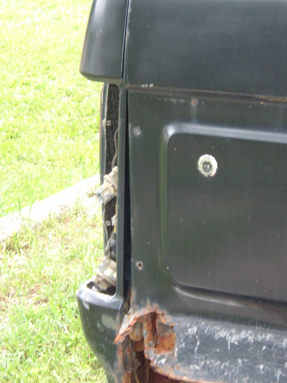 Rear corner of a dark-colored vehicle showing a damaged taillight area and significant rust on the bumper