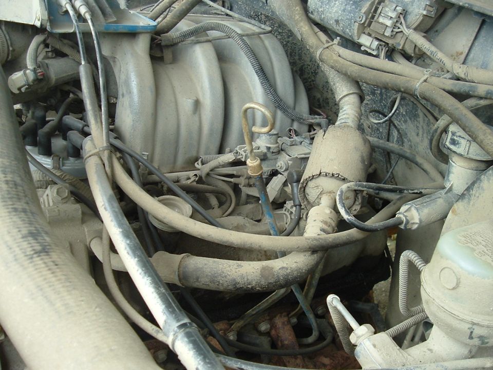 Engine bay with dusty intake manifold, spark plug wires, and hoses. Some text visible on a white container