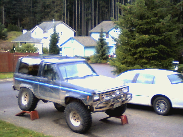 Lifted blue Ford Bronco II on ramps in a driveway with big off-road tires and brush guard, white sedan beside it