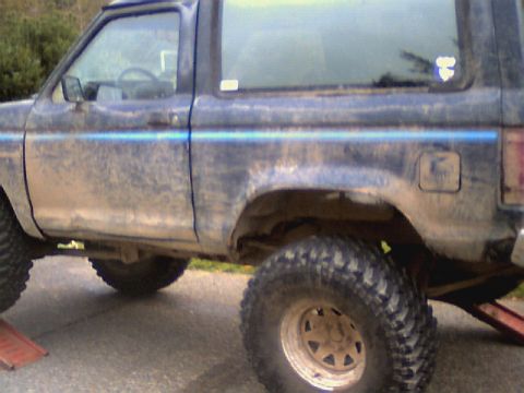 1987 Ford Bronco II - This lifted SUV, possibly a Ford Bronco II, is shown from the side with large off-road tires and a distinctive blue stripe.