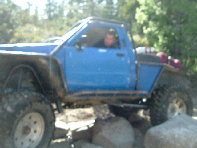 Blue solid-axle rock crawler built from a small pickup cab climbing over boulders, driver inside