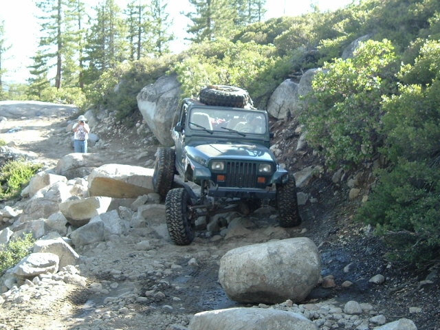 Dark green Jeep Wrangler YJ with rooftop spare climbing a steep rocky trail, person watching at left