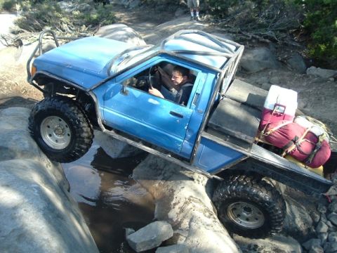 1989 Ford Bronco - A heavily modified blue pickup truck tackles a rocky trail with a driver inside and camping gear in the bed.