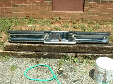 1985 Ford Bronco - A chrome rear step bumper with black rub-strip top and a bolt-on receiver/hitch plate with ball rests upright on a landscape timber against…