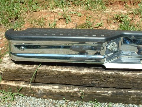 1985 Ford Bronco - A chrome rear bumper with a black step pad is shown, resting on weathered wooden beams outdoors.