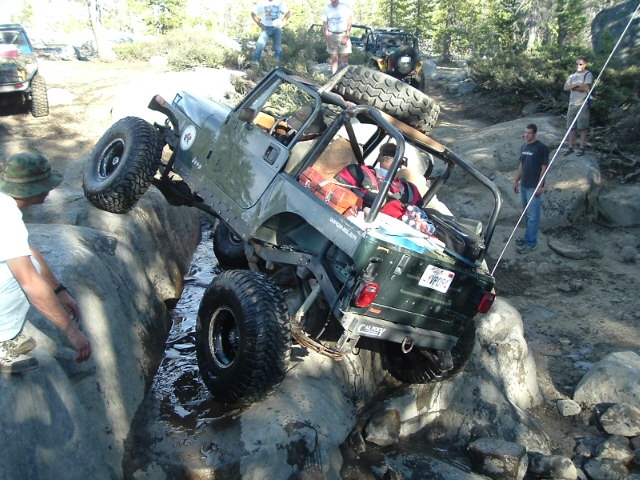 Olive-green Jeep 4x4 crawling wet granite boulders as onlookers watch