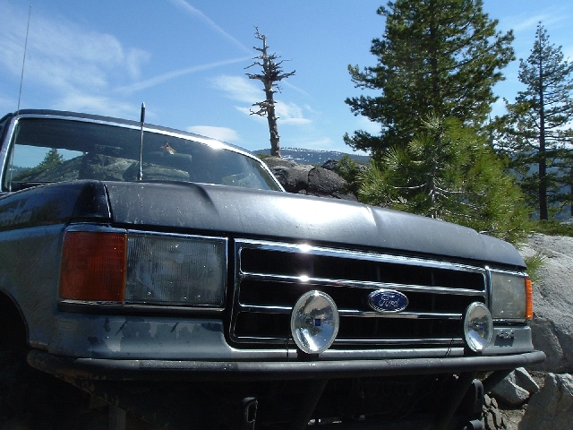 Front end of dark Ford pickup with chrome grille and Ford oval, parked on rocky high-country trail