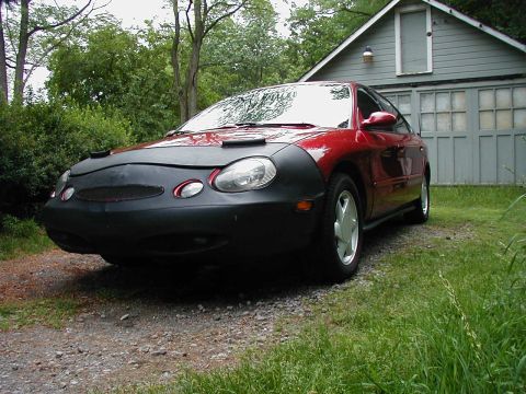 1997 Ford Taurus - A red sedan is parked in front of a garage, partially covered by a black car bra.