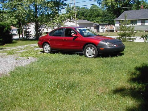 1997 Ford Taurus - A red sedan sits in a grassy yard on a sunny day. The car has a black bra covering the front bumper and hood.