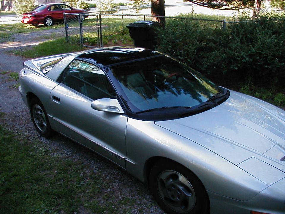 Silver Pontiac Firebird with a black roof, parked on gravel. A red sedan is in the background