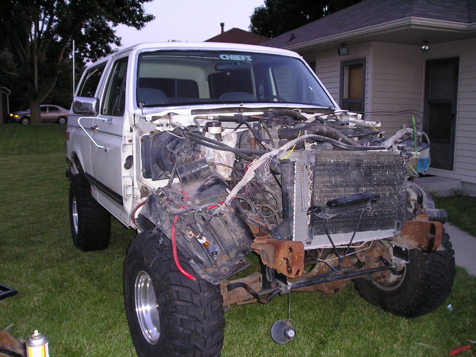 Lifted white Bronco with front clip off and radiator core hanging out, parked on lawn by a house