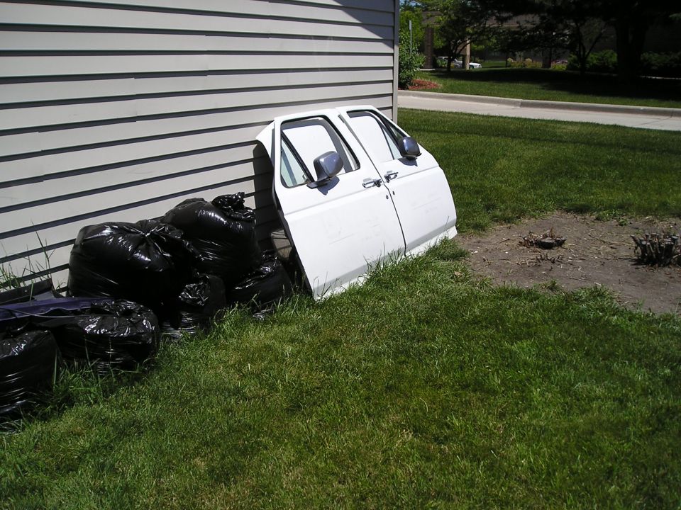 White truck door leaning against house siding next to filled black trash bags on the lawn
