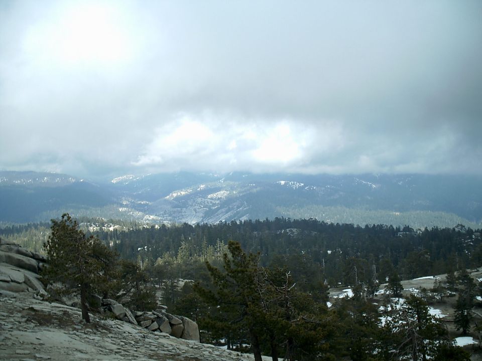 Mountain landscape with pine trees, rocks, and snow patches under a cloudy sky. Supermotors.net logo in lower right