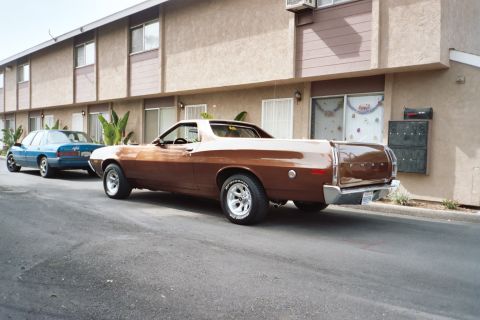 1972 Ford Ranchero - A brown 1972 Ford Ranchero is parked next to a blue sedan in front of an apartment building.
