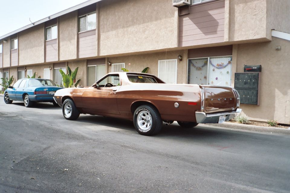 Brown Ford Ranchero pickup parked at curb, rear 3/4 view, with blue sedan behind