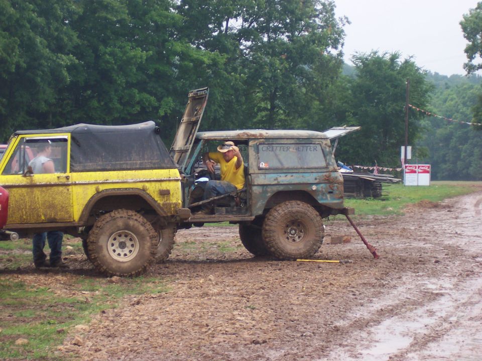 Yellow early Bronco and a green flatbed 4x4 parked in a muddy field at an off-road event
