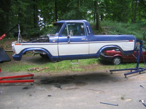 1978 Ford Bronco - Blue and white two-tone Ford Bronco body shell lifted off its frame and resting on stands in a wooded driveway, with the hood, doors, roof,…