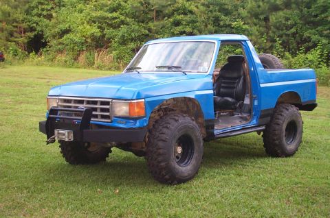 1987 Ford Bronco - This lifted pickup truck, likely from the early 1980s, is parked on a grassy field with trees in the background.