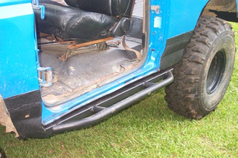 1987 Ford Bronco - Aftermarket rock sliders are visible on the side of a blue vehicle, with a view of the interior floor and a large tire.