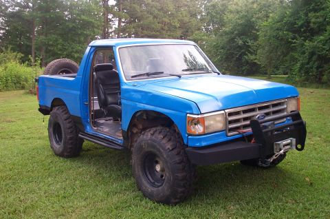 1987 Ford Bronco - This lifted 1987 Ford Bronco sits on a grassy lawn with trees in the background. The driver's side door is open, revealing the interior.