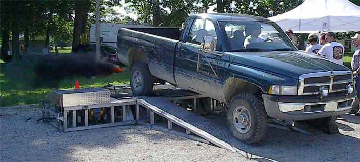 Dark green Dodge Ram pickup on a portable dyno, black smoke billowing, at an outdoor event